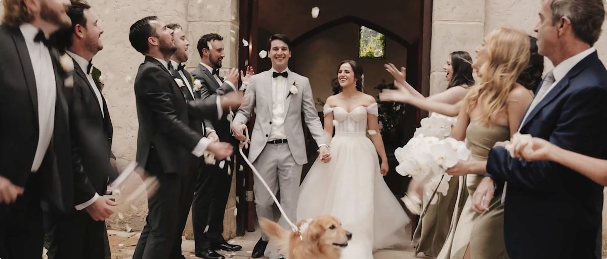 A wedding party throws confetti over a couple leaving a stone chapel, with a golden retriever standing among the guests.