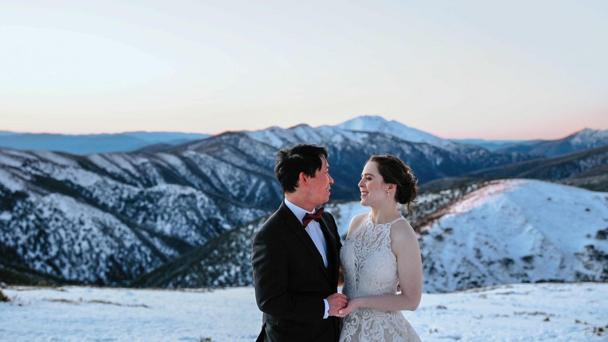 A couple in wedding clothes stand together on a snow-covered alpine ridge.