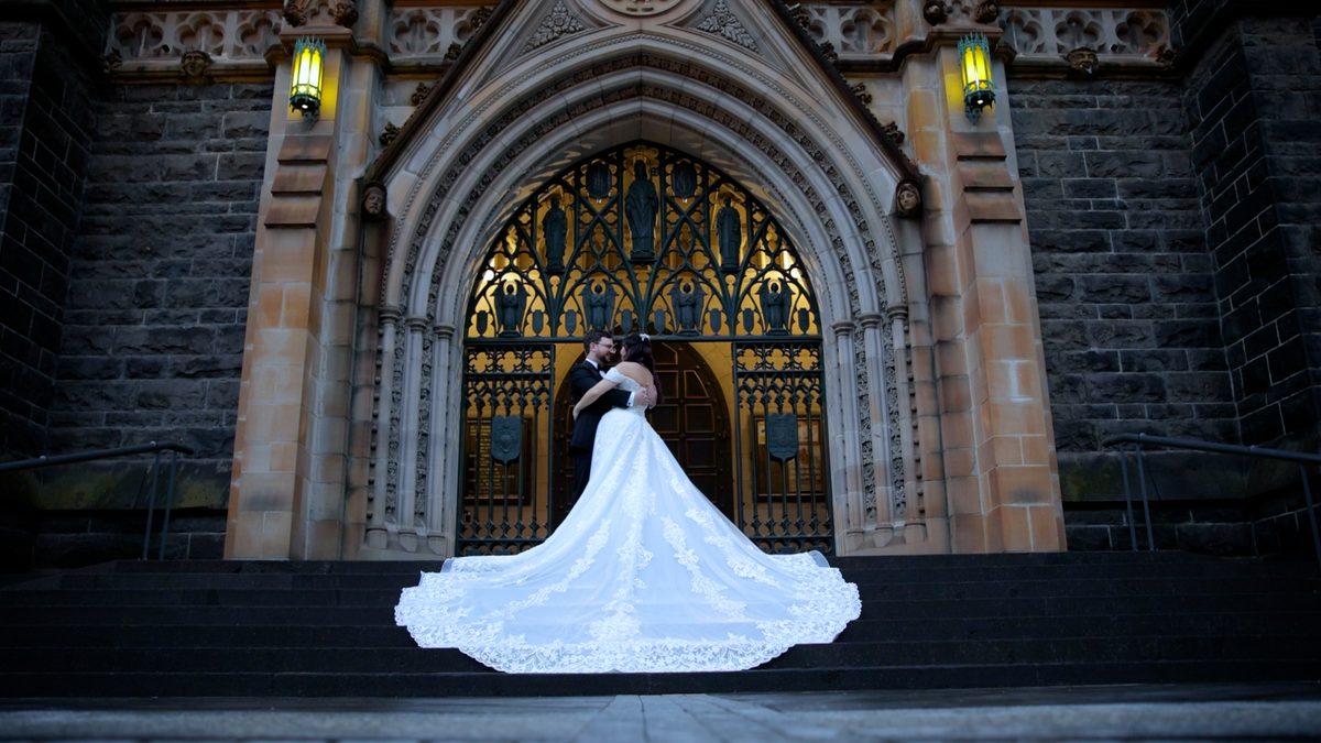 A bride stands in the arched doorway of a bluestone cathedral at night, her long train spread down the steps behind her.
