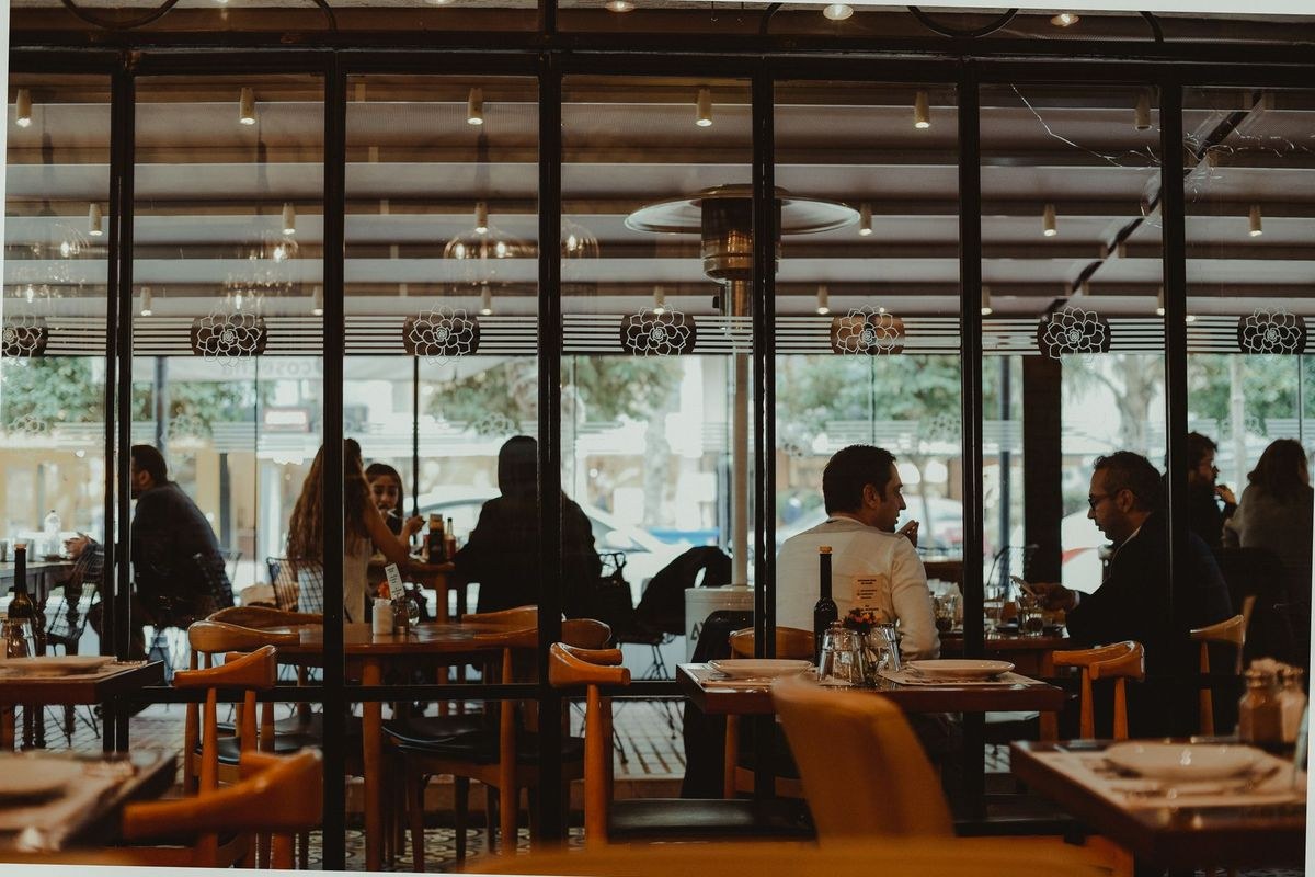 Diners at timber tables in a warm, street-facing restaurant dining room.