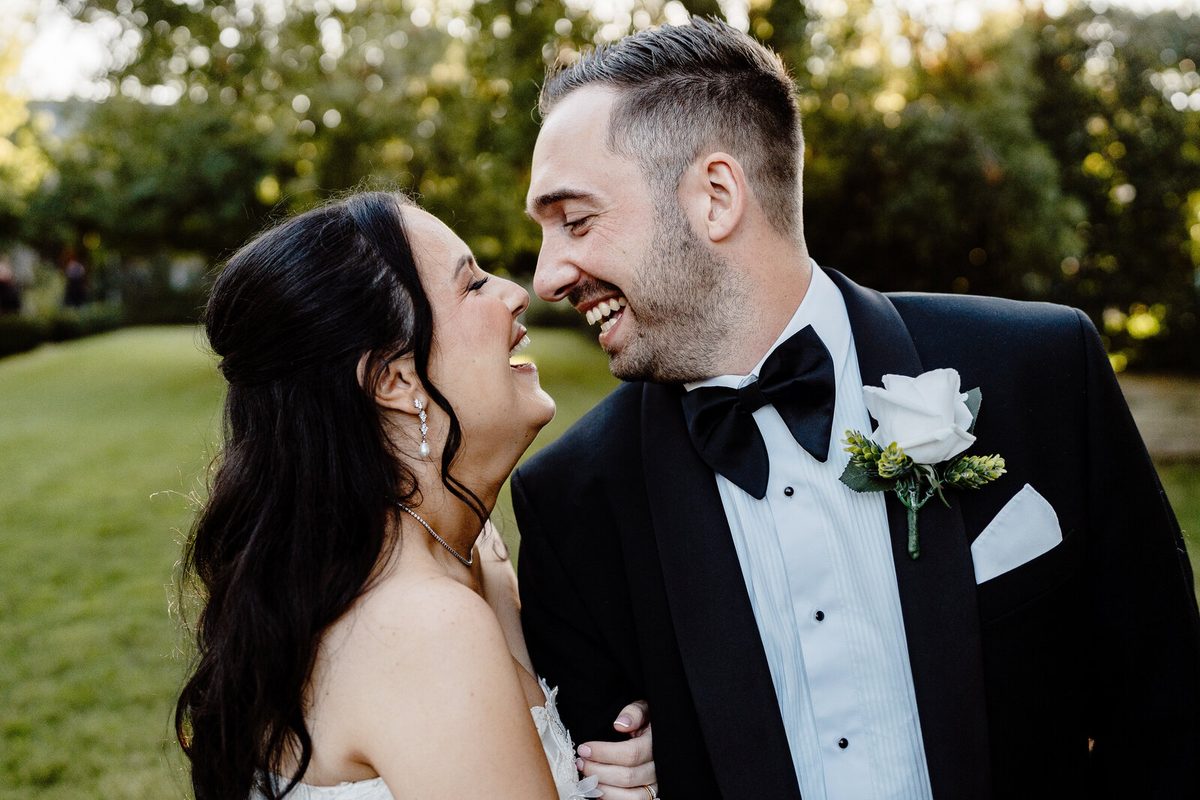 A bride and groom laugh together in close-up on a lawn, foreheads almost touching.