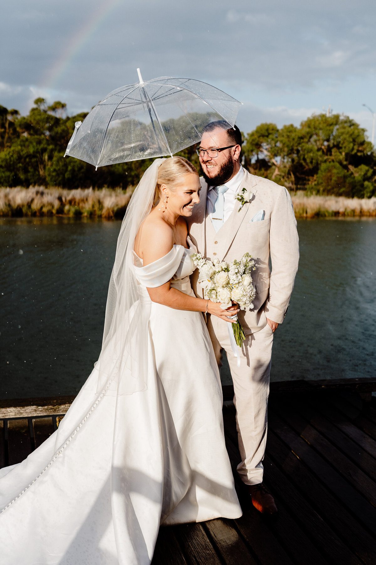A couple stand under a clear umbrella beside a lake, laughing, with a faint rainbow in the sky behind them.