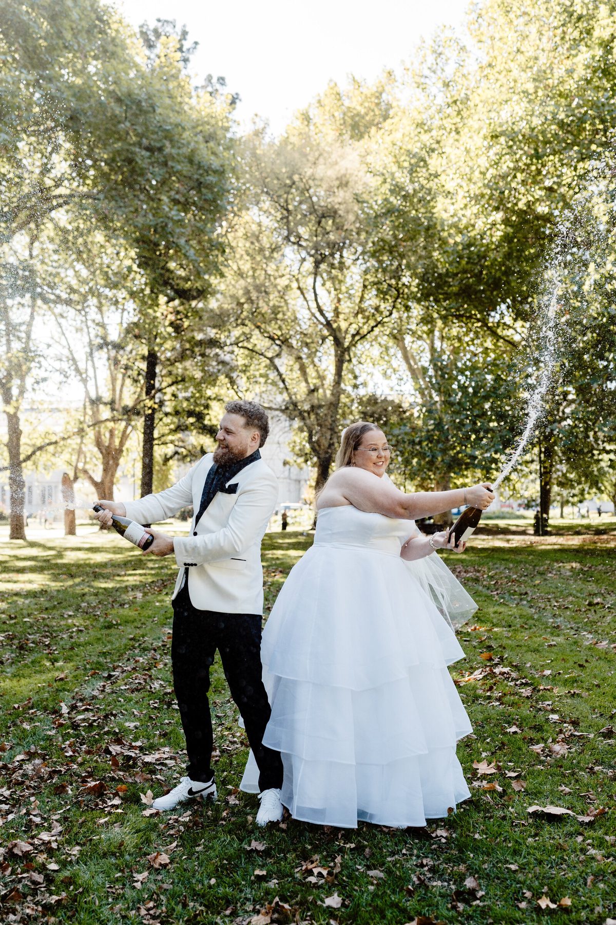 A couple spray a bottle of champagne on a lawn, both laughing, with the spray arcing through sunlight.
