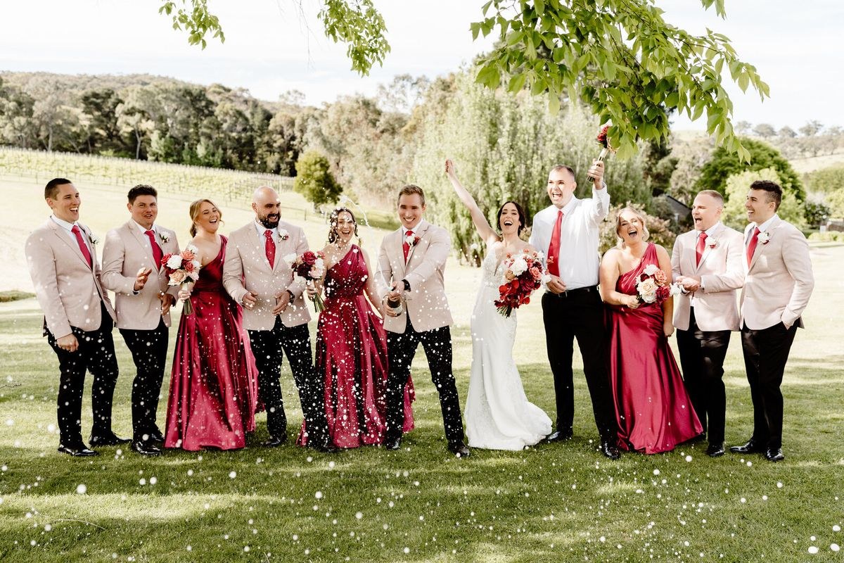 A bridal party in cream suits and deep red dresses laughing as confetti falls around them on a vineyard lawn.