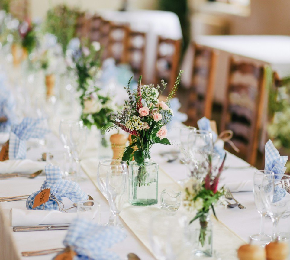 A long reception table set with gingham napkins, posies in glass jars and wine glasses.