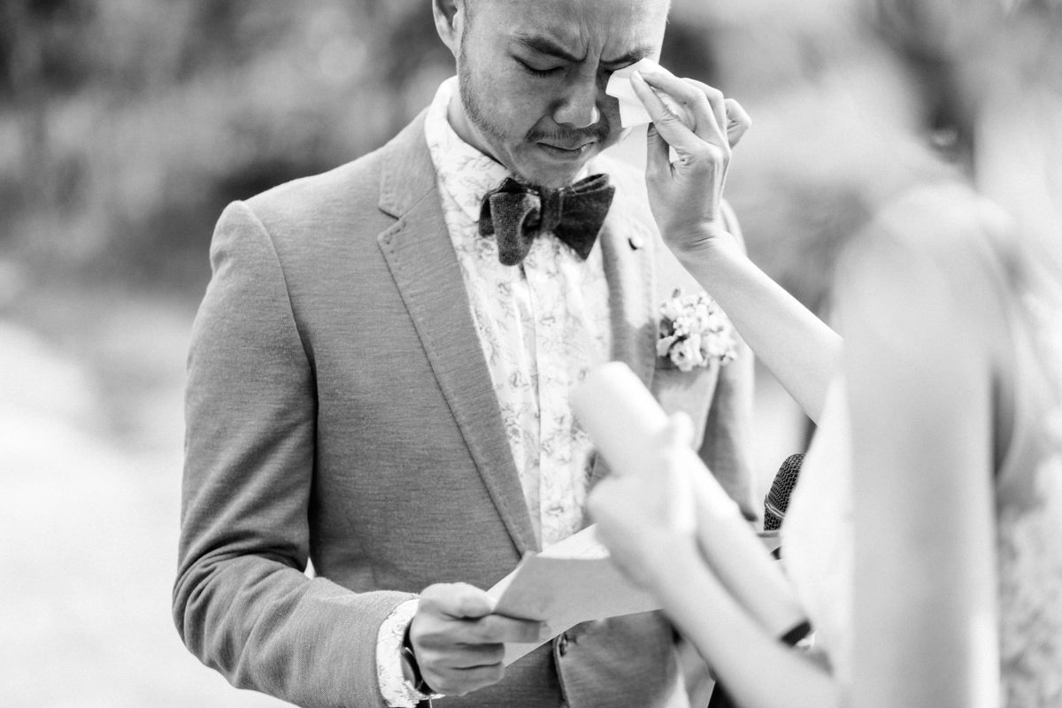 A black and white photograph of a groom wiping his eye with a handkerchief while reading from a piece of paper.