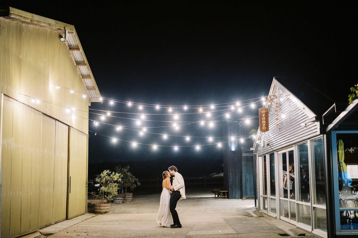 A couple dance alone at night on a lit forecourt beside a barn, under strings of festoon lights.