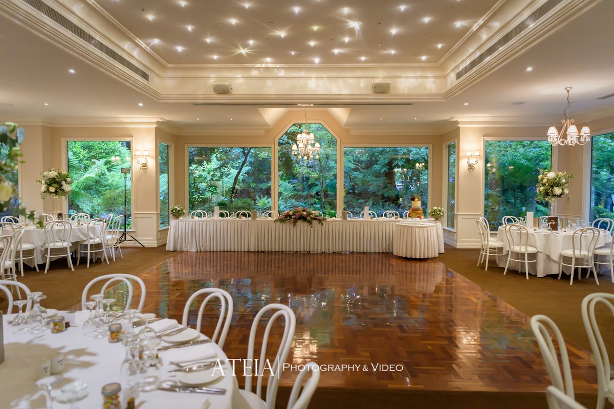 A reception room with a long bridal table, round guest tables and a parquet dance floor, with forest visible through tall windows.