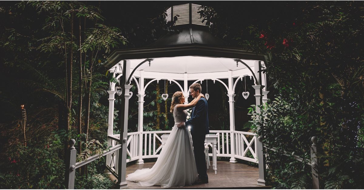A couple stand inside a white lit gazebo surrounded by dense forest at night.