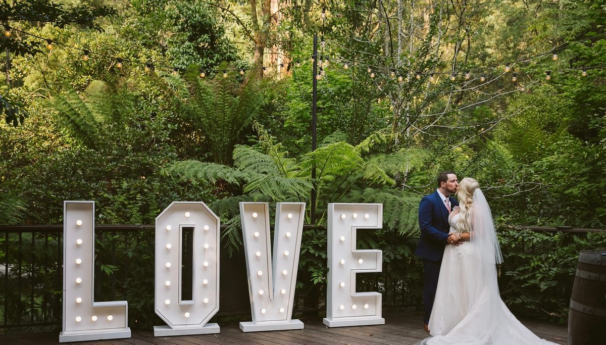 A couple embrace on a deck at dusk beside illuminated LOVE letters under strings of festoon lights.