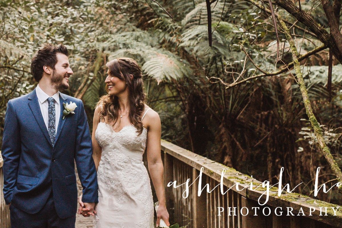 A couple walk hand in hand across a timber bridge surrounded by tree ferns in the Dandenong Ranges.