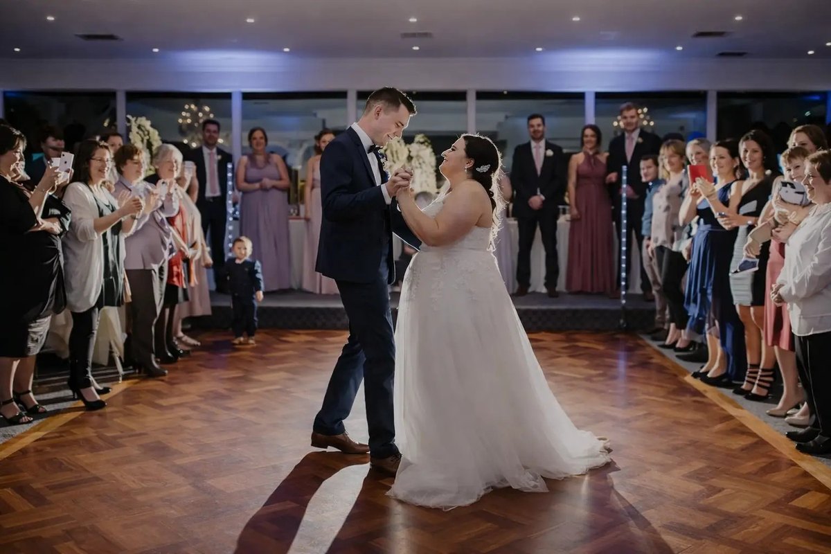A couple's first dance on a parquet floor under purple uplighting, with guests ringing the dance floor.
