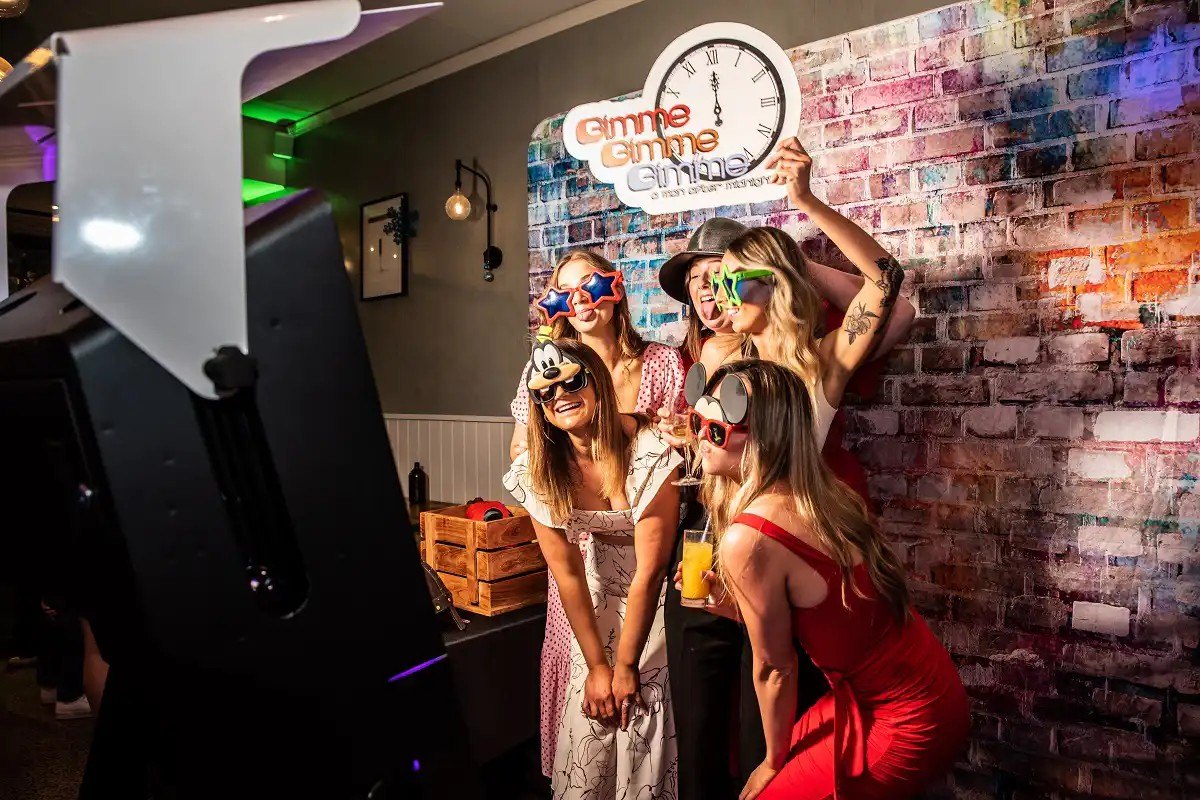 Five guests in novelty glasses and props pose in front of a colourful brick backdrop beside the mirror booth.