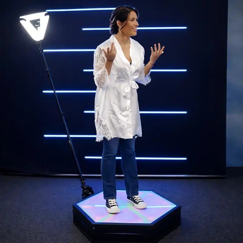 A guest in a white robe stands on a lit hexagonal 360 booth platform with a ring light on an arm beside her.