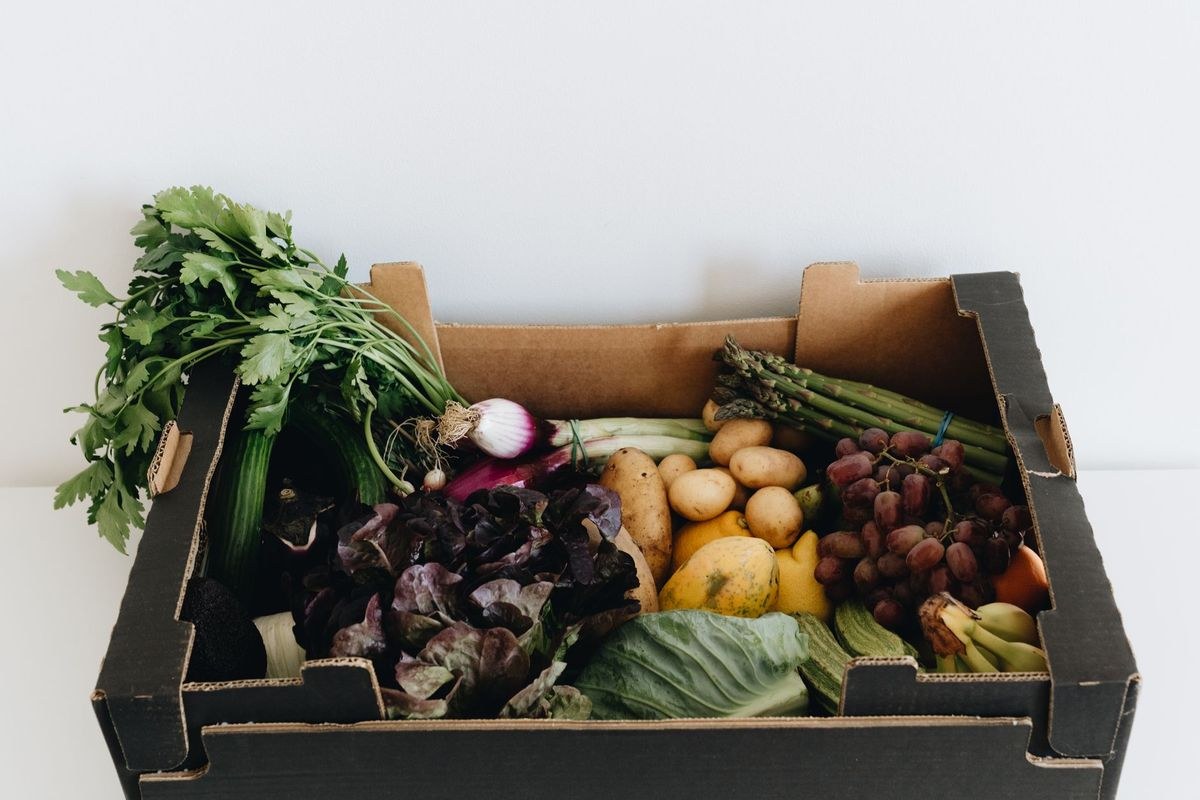 A cardboard box packed with fresh produce — leafy greens, potatoes, grapes, asparagus and herbs — ready for redistribution.