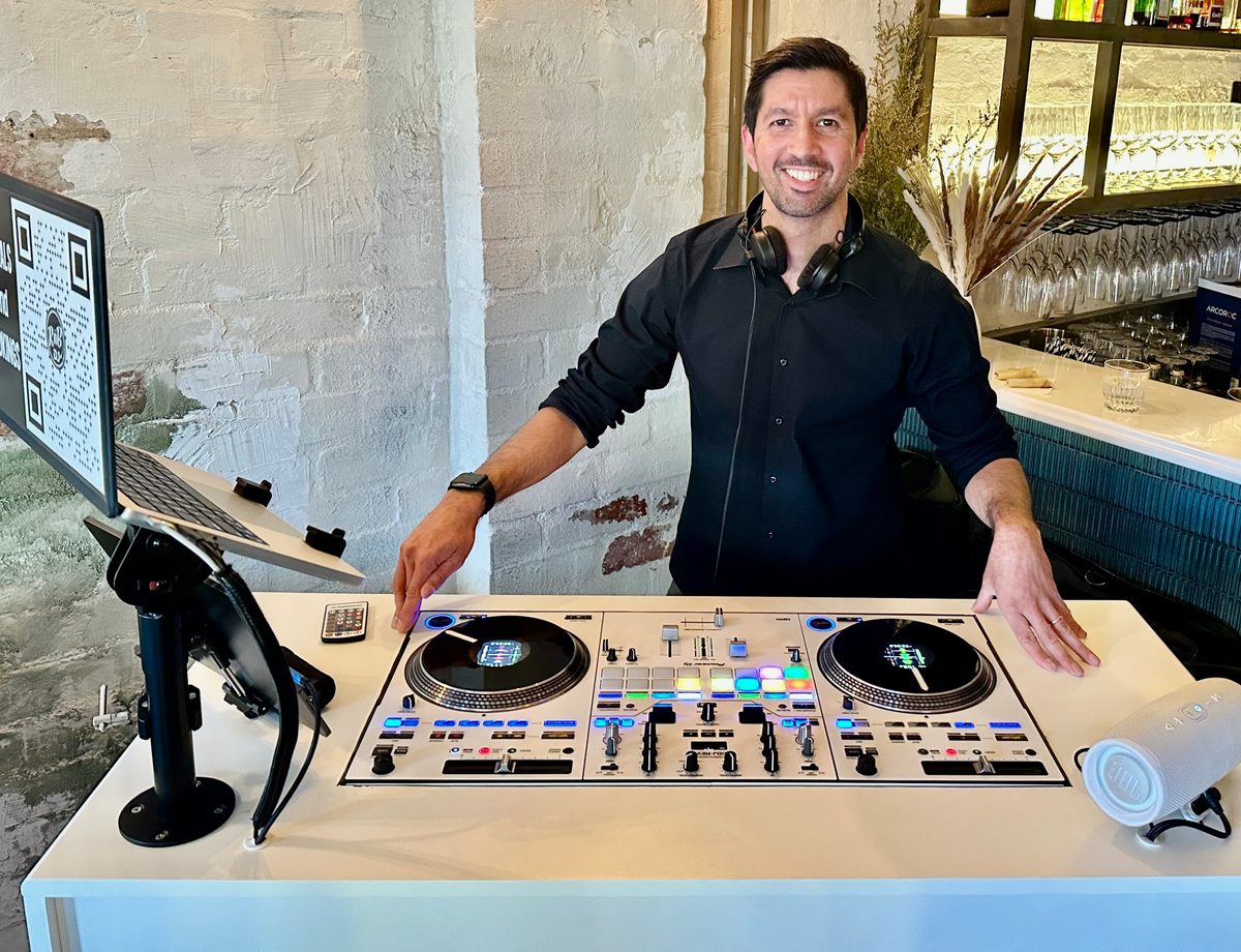 A DJ in a black shirt stands behind a white booth with a controller, turntables and a laptop at a function venue.
