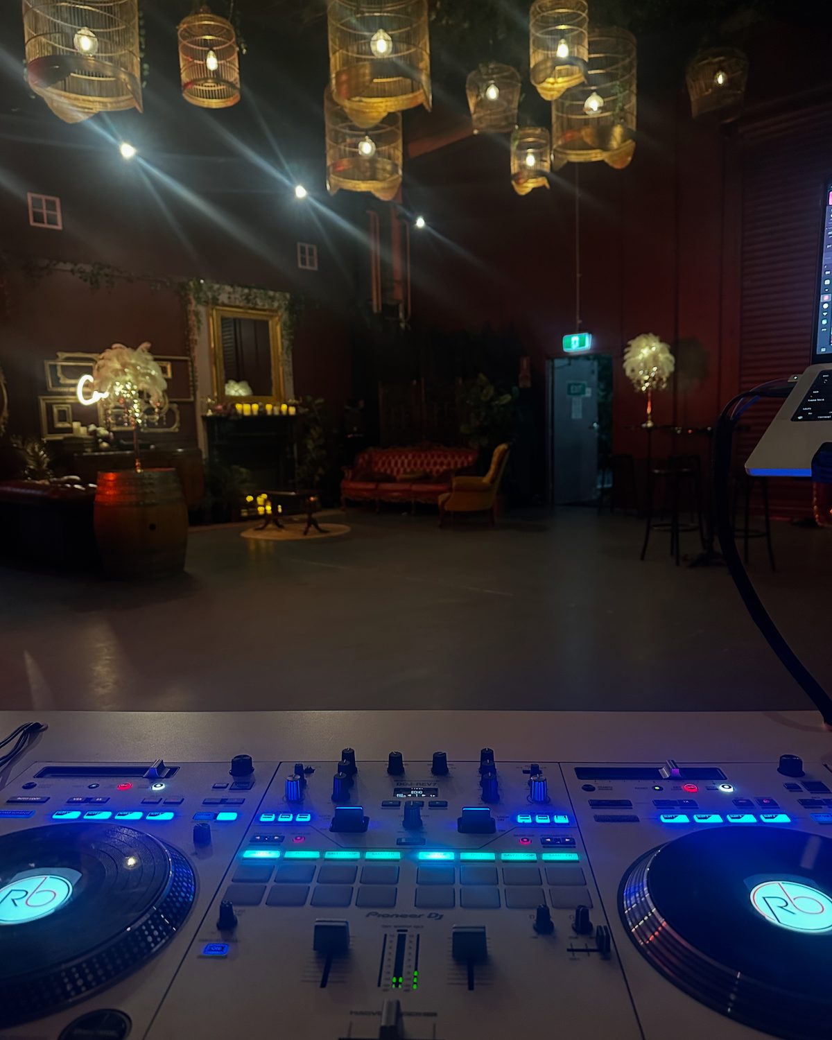 A lit DJ controller in the foreground with an empty candlelit function room and hanging lanterns behind it.