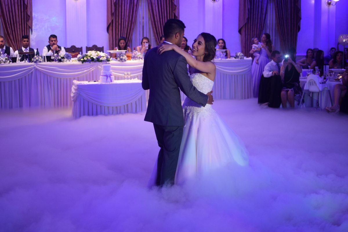 A couple dance in thick low fog under purple uplighting while seated guests watch from tables behind them.