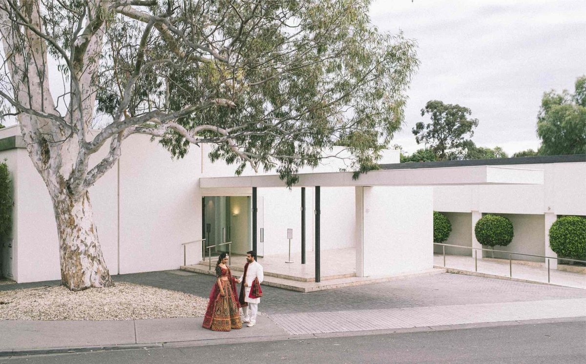 A couple in Indian wedding dress stand outside a low white modernist building, framed by the trunk of a gum tree.