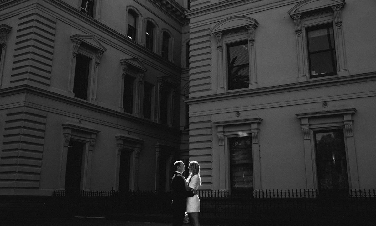 A black and white photograph of a couple standing small in the doorway of a grand bluestone building.