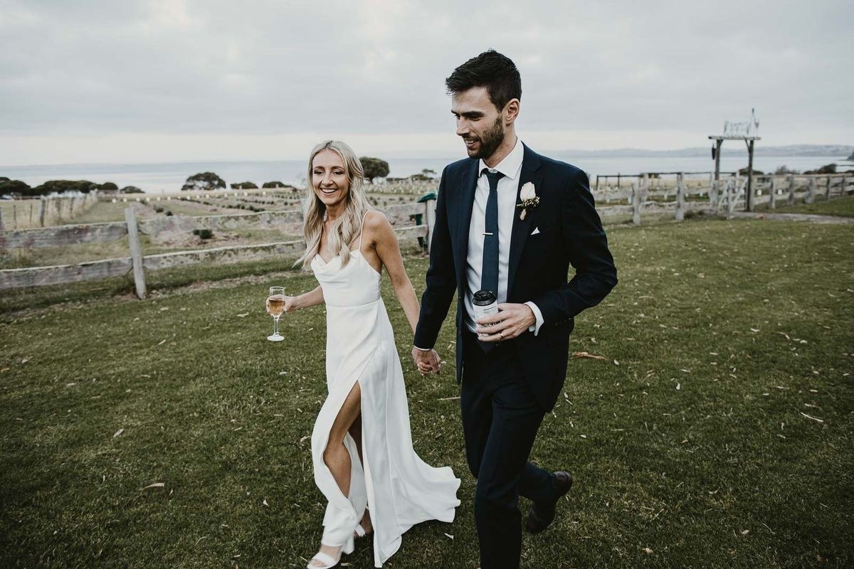 A couple walk across grass with drinks in hand, vineyard rows and the coastline behind them under a grey sky.