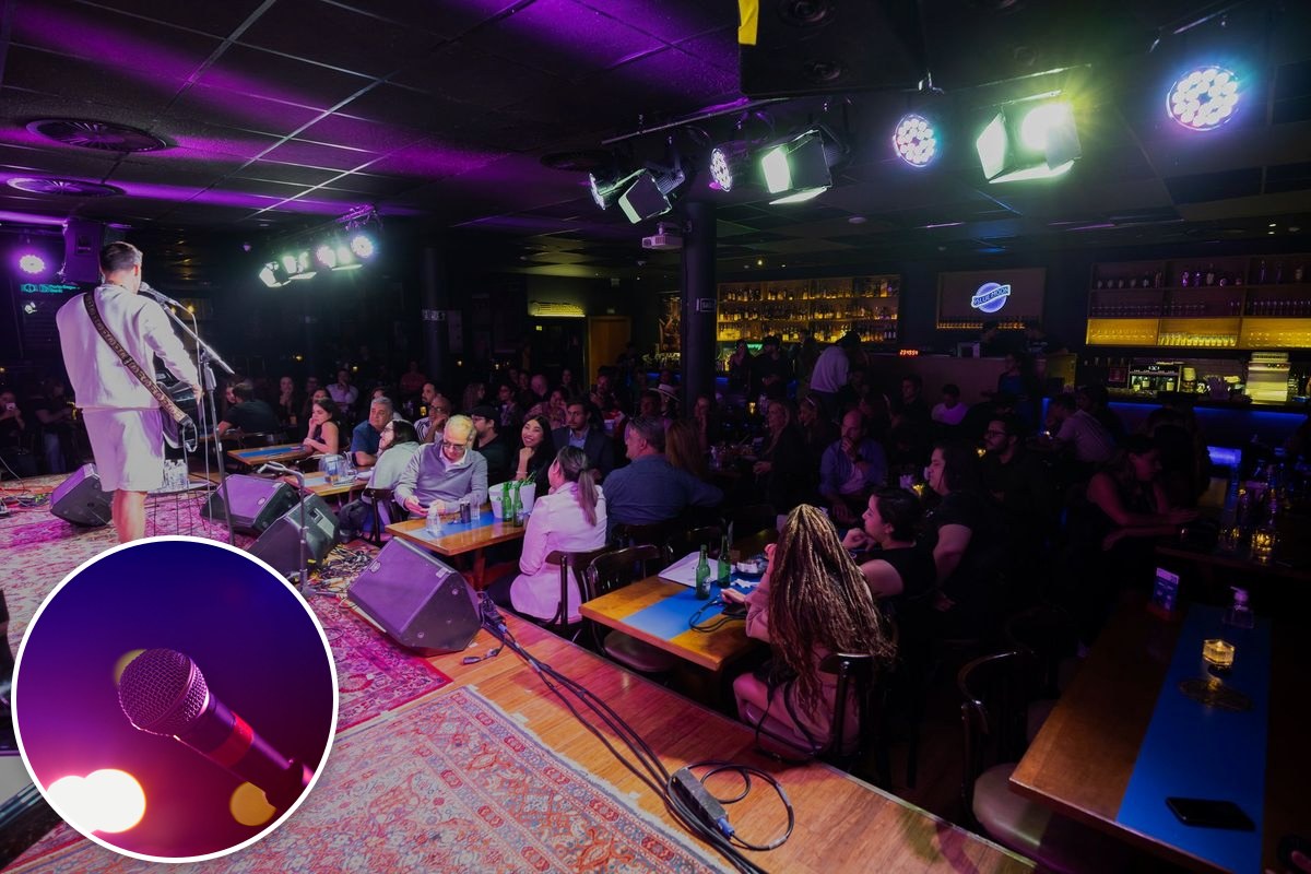 A musician performs on a small stage in a busy, dimly lit club as a seated crowd watches.